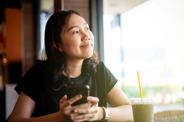 Asian ethnicity young woman relaxing and drinking iced coffee at coffee shop. Woman using a smartphone to send message while drinking a coffee in cafe and restaurant close up with copyspace.