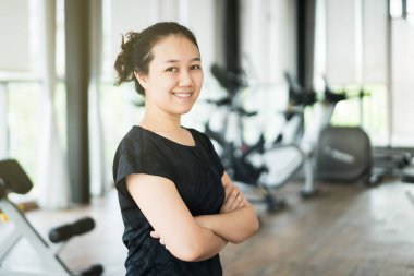 Spor salonunda ağırlık antrenmanı ve kardiyo egzersizinden sonra fotoğrafçılık için poz veren ve gülümseyen Asyalı güzel bir spor kadını. Spor salonunda vücut çalışan sağlıklı zayıf bir kadın. Arka planda fotokopi makinesi olan egzersiz makineleri..