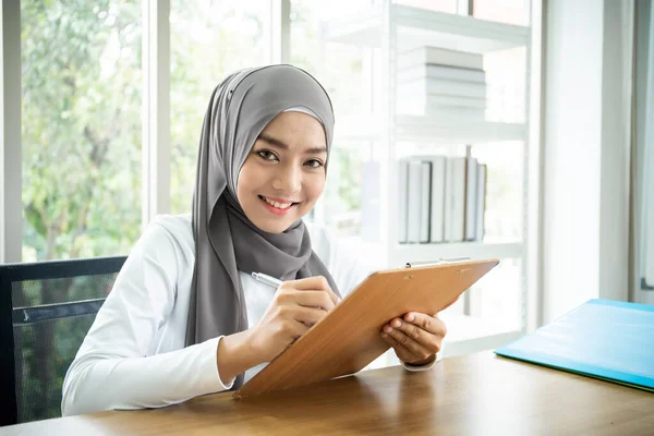 Asian muslim businesswoman writing on a clipboard. Multicultural and diverse ethnicity of people in workplace. Muslim secretary prepare a document for her boss. Modern muslim businesspeople concept.