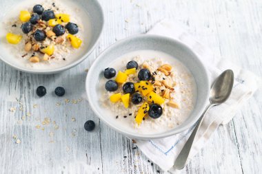 Mangolu yulaf lapası, yaban mersini ve beyaz ahşap masada doğranmış badem. Seçici odaklanma. Açık arkaplanda parlak, sağlıklı kahvaltı görüntüsü, mesaj için yer. Süperyiyecekler.