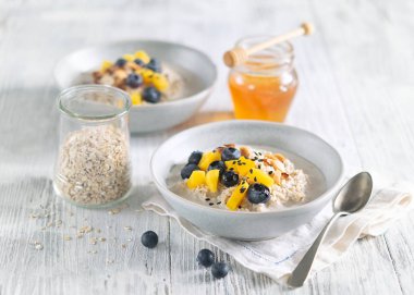 Mangolu yulaf lapası, yaban mersini ve beyaz ahşap masada doğranmış badem. Arka planda bal kavanozu. Seçici odaklanma. Açık arkaplanda parlak, sağlıklı kahvaltı görüntüsü, metin için boşluk.