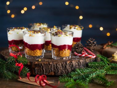 Krem peynir, kırmızı meyve sosu, kurabiye kırıntıları koyu mavi arka planda bardakta tatlı, çelenk ışıkları bokeh, Noel süslemesi. Yeni yıl, Noel tatili konsepti.
