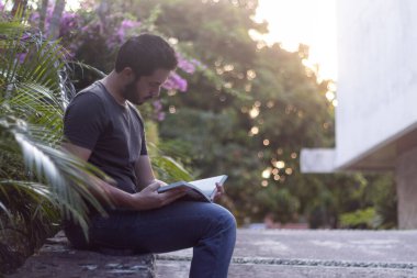 Duvarda oturan genç adam, gün batımında kitap okurken elinde tutuyor.