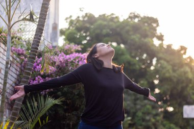 Bahçede genç bir kadın, kolları açık özgürlük ve sükuneti ifade ediyor.