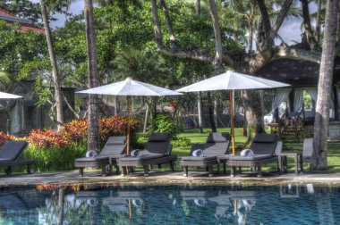 Yüzme havuzunun yanındaki büyük şemsiyelerin altında uzanan sandalyeler. 