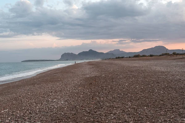 Gün batımında sahilde, dağların önünde. Kızıl gökyüzünde bulutlar olan sakin bir deniz manzarası..