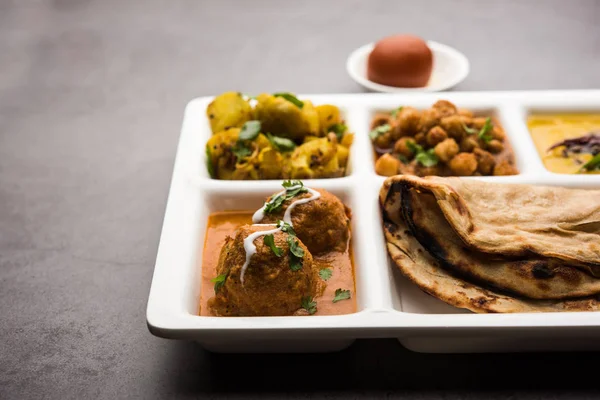 Indian vegetarian Food Thali or Parcel food-tray with compartments in which Malai Kofta, chole, Dal tarka, dry aloo sabji, chapati and rice with sweet gulab jamun served