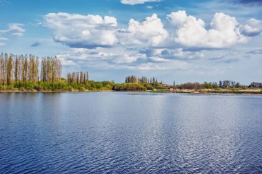 Nehir kıyısında güneşli bir gün - kıyı şeridi, ağaçlar ve mavi gökyüzü suya yansıyor.