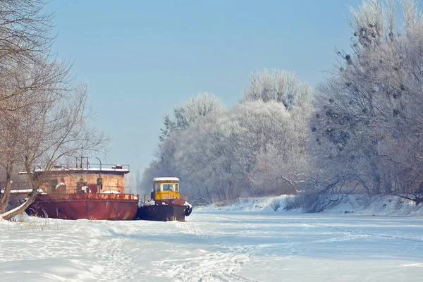 Winter landscape - frozen river with tugboat and barge, trees on the banks.