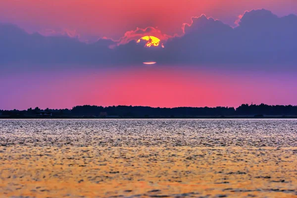 Big sun in clouds over silhouette of lake shore at sunset.