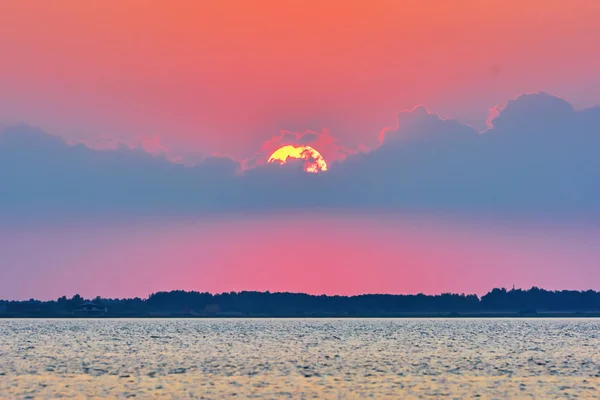 Big sun in clouds over silhouette of lake shore at sunset.