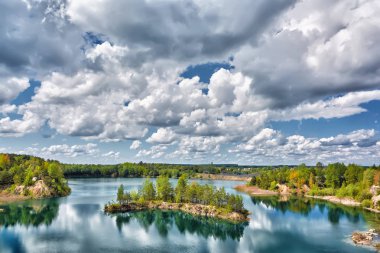 Ada ile göl ve ormanla kaplı kayalık kıyılar. Mavi gökyüzü suya yansıyan bulutlarla.
