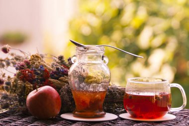 Glass cup of hot herbal tea, a small jar of jam with a teaspoon and a red apple beside a dried flower bunch on a blurred colorful background
