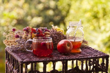 Bir bardak sağlıklı bitki çayı, bir çay kaşığı reçel ve kırmızı elma ile bulanık yeşil ve sarı arka planda kurumuş bir demet çiçeğin yanında