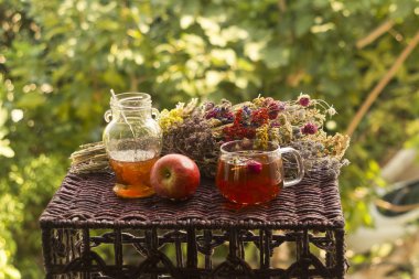 Bir bardak sağlıklı bitkisel çay, küçük bir kavanoz reçel ve kırmızı elma. Renkli, kurutulmuş bir bitki ve çiçeğin yanında.  