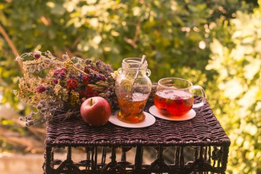 Bir bardak sağlıklı bitkisel çay, küçük bir kavanoz reçel ve kırmızı elma. Renkli, kurutulmuş bir bitki ve çiçeğin yanında.  