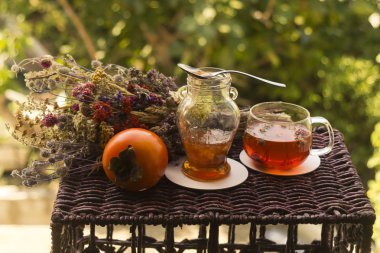 Bir bardak sıcak bitkisel çay, küçük bir kavanoz reçel ve turuncu bir hurma meyvesi. Renkli, kurutulmuş bir bitki ve çiçek tomarının yanında.  