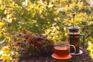 Bir bardak sıcak, sağlıklı bitki çayı, küçük bir çaydanlık ve renkli, kurutulmuş bir bitki ve çiçekli demet güneş ışığıyla aydınlanan bahçede küçük bir rattan masasında.