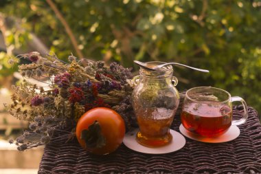 Bir bardak sıcak bitkisel çay, küçük bir kavanoz reçel, bir portakallı hurma meyvesi ve bahçe masasında renkli kurutulmuş bitki ve çiçek demeti. 