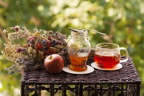 Bir bardak sıcak bitkisel çay, bir çay kaşığı reçel, bir kırmızı elma ve bahçedeki küçük bir rattan masasında renkli kurutulmuş bitki ve çiçek demeti.  