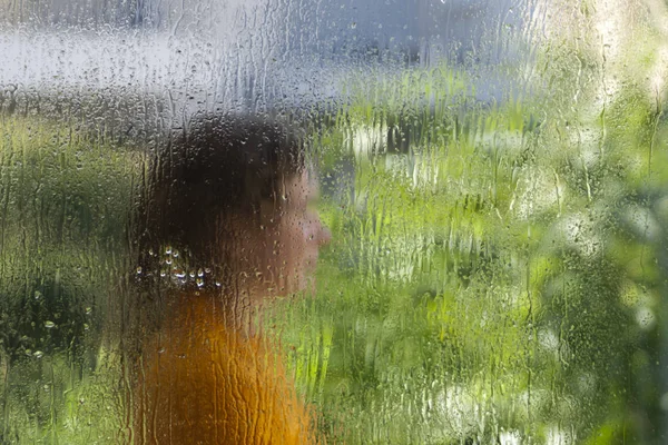 Wet window glass with a defocused silhouette of a woman in an orange dress and blurred green leaves