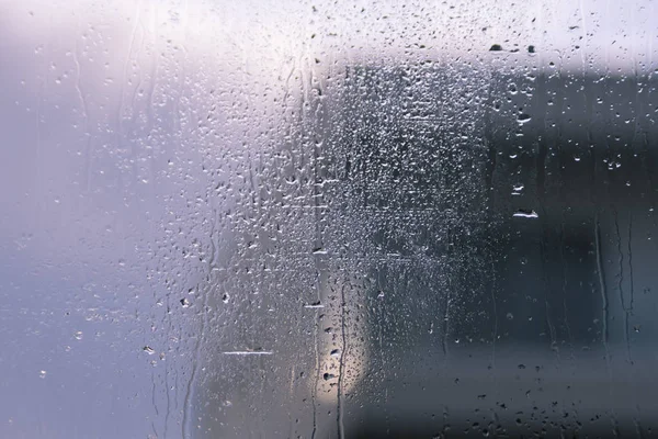 Wet window glass with lots of rain drops and a view of the pale sky and a gray silhouette of a house