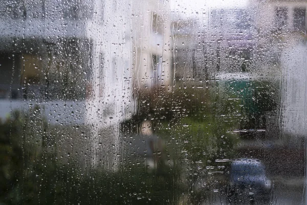Wet window glass with lots of rain drops and a view of the blurred silhouettes of houses, trees and a car on the street