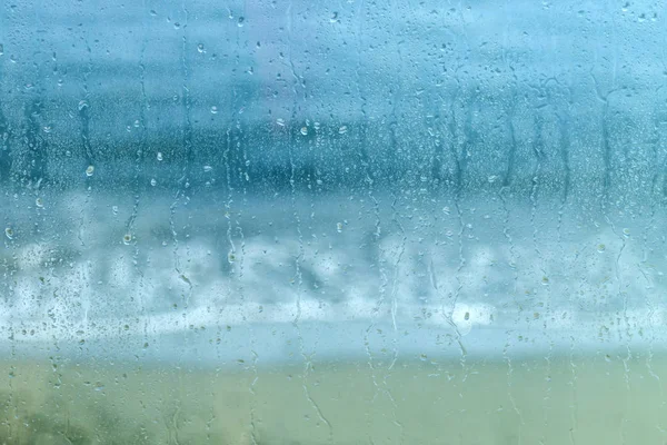 Wet window glass with water drops and a blurred view of the blue sea and yellow sand at the beach