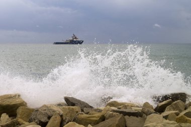 Beyaz deniz dalgası kara bulutlu gökyüzü ve ufukta bir gemi ile kayalara çarpıyor.