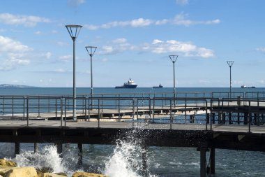 Sokak lambaları, mavi deniz, bulutlu gökyüzü ve ufukta üç gemi olan boş bir rıhtım manzarası