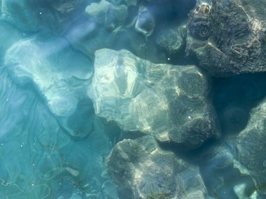 Şeffaf turkuaz deniz suyu yüzeyinin ve suyun altındaki kayaların yakın çekimi.