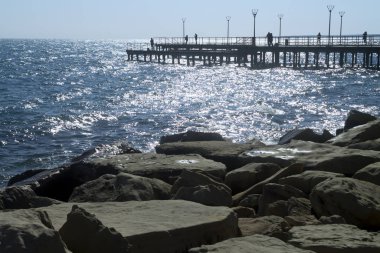 Rocky coastline, glittering sea and a pier with street lamps and silhouettes