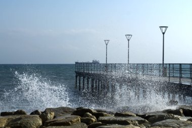 Empty sea pier with street lamps, splashing waves, the blue sky and a ship on the horizon