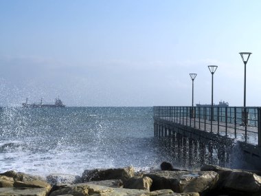 Kayalara vuran dalgalar, ufukta gemileri olan deniz, açık mavi gökyüzü ve sokak lambaları olan boş bir iskele.