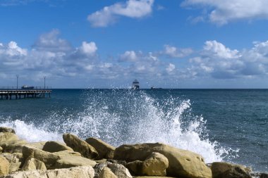 Su sıçratan dalgalı kayalar, ufukta gemileri olan bir deniz manzarası ve gri bulutlu mavi gökyüzü.