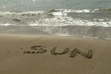 Kelime Güneşi sahildeki ıslak kumda yazılıydı. Deniz gün ışığında parlıyordu.