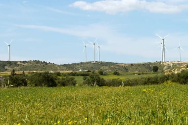Tarım tarlaları, yeşil tepeler, rüzgar türbini kuleleri ve yukarıdaki mavi gökyüzü ile kırsal alan.