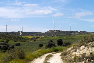 Kırsal bir arazi, tarımsal tarlalar, yeşil tepelerdeki rüzgar türbini kuleleri ve beyaz bulutlu mavi gökyüzü.