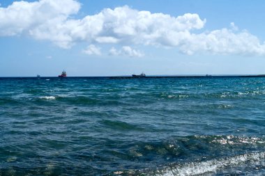 Dalgalı mavi deniz suyu yüzeyi, ufukta gemileri olan beyaz bulutlu gökyüzü.