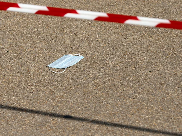 Used disposable medical face mask lying on the road with red and white striped barricade tape