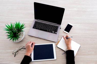 Kadın, laboratuvar, tablet ve akıllı telefon gibi çeşitli ofis aletleriyle çalışıyor. Ofisteki ahşap masanın üzerindeki küçük deftere yazıyor..