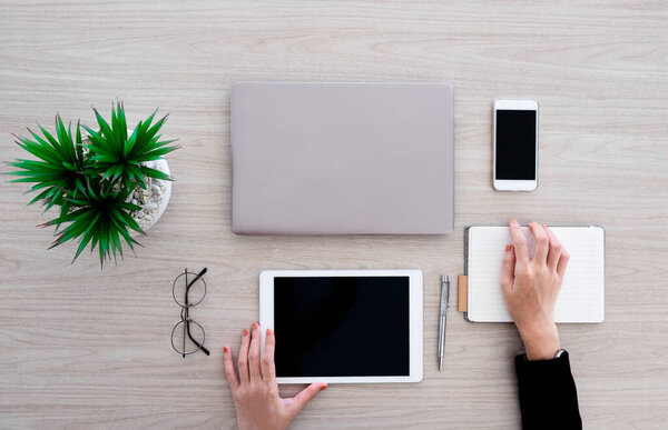 The woman is working with various office gadgets like labtop, tablet and smartphone. She is  writing on the small notebook  on the wooden desk in office.