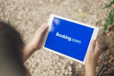 RAYONG, THAILAND, APR 12.2020: Booking.com uygulaması üzerinden seyahat bilgilerini aramak için ekranda Apple Ipad 'i Booking.com logosuyla tutan Asyalı adam.