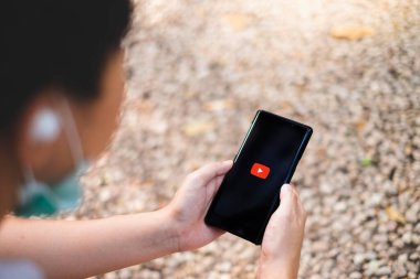 RAYYONG, THAILAND, APR 12.2020: Karantina zamanı arasında eğlence için mobil ekranda logolu Youtube uygulaması kullanan bir adam. Ayrıca cerrahi maske ve kulaklık takıyor..