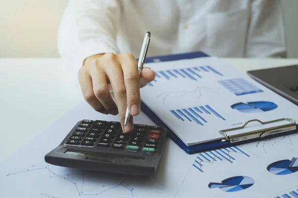 Ofisteki beyaz masadaki grafik grafiğini ve iş verilerini hesaplamak için elinde kalem tutan ve hesap makinesi kullanan işadamının yakın plan çekimi. finans muhasebe kavramı.