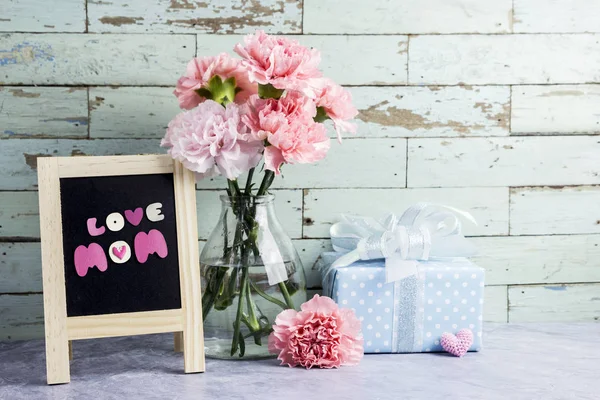 Mothers day concept of pink carnation flowers in clear bottle and gift box and love mom text on blackboard — Stock Photo, Image