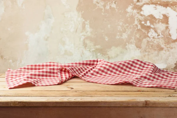 Empty wooden table with red checked tablecloth over rustic wall background.