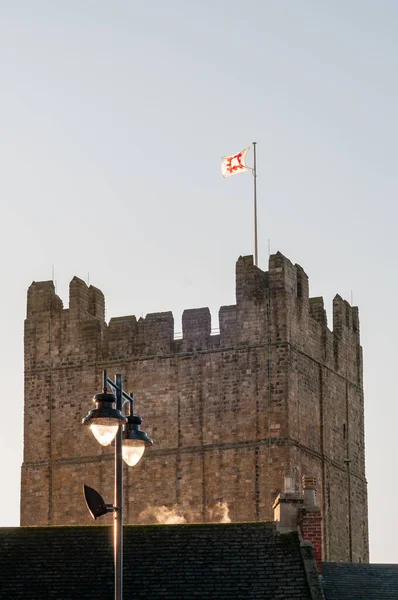 English Heritage flag atop the keep of Richmond Castle, North Yorkshire with steam rising from rooftops in the foreground and catching the golden, early morning sunlight