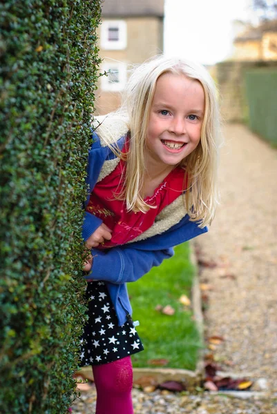 Pretty young blonde girl peeks out from behind a hedge — Stock Photo, Image