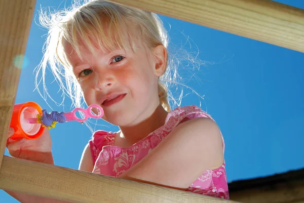 A Closeup of a pretty, young girl wearing a pink dress, about to blow bubbles from the top of her climbing frame, on a sunny summers day with a deep blue sky — Stock Photo, Image